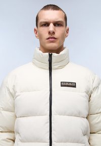 Gevoerde crème jas met hoge kraag, zwarte rits en logo patch op de borst. Heeft horizontale gestikte secties voor isolatie.