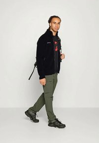 Mustavilline tõmblukuga fliisjakker, millel on logo, koos punase graafikaga t-särgiga ja roheliste cargo-pükstega. Mustad sportjalatsid lisavad kontrasti.