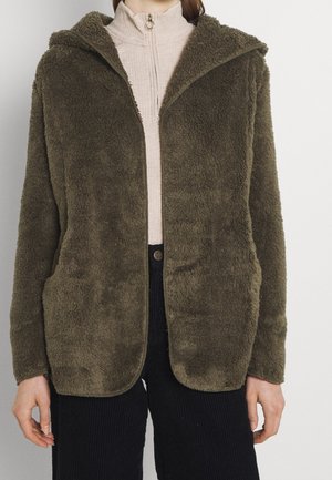 Personne portant une veste duveteuse vert olive ouverte sur un haut beige zippé et un pantalon noir sur fond uni.