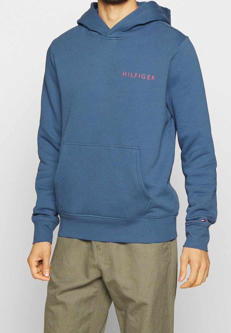 Felpa blu con tasca a canguro frontale, caratterizzata da un piccolo logo rosso "HILFIGER" sul petto. Tessuto in cotone misto con una consistenza morbida.