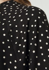 Černá látka s bílými puntíky, která se vyznačuje měkkou strukturou a stylovým vzorem. Látka se hladce splétá, což umocňuje celkový design.