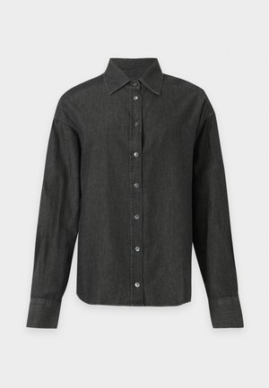 Jeune femme aux cheveux courts et foncés portant une chemise noire ample à boutons, un jean gris et des bottines noires sur un fond clair uni.