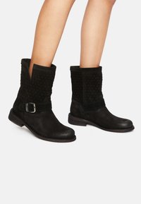 Black suede ankle boots feature a textured upper, flat heel, and a side strap with a silver buckle. Cut-out detail at the top.