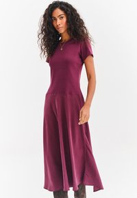 Bordowa sukienka z dopasowaną górą i rozkloszowanym dołem. Krótkie rękawy, miękka tekstura, sięga do połowy łydki. Minimalistyczny design, bez widocznych wzorów czy zdobień.