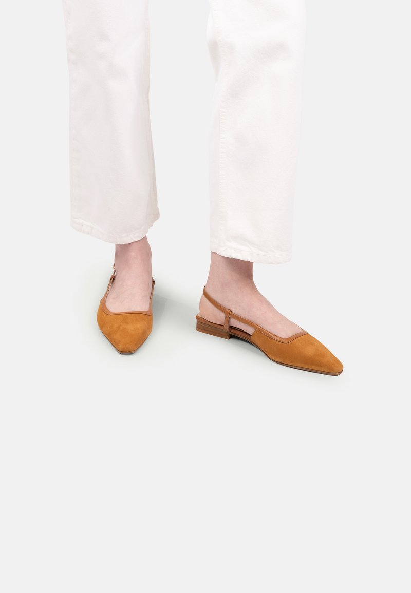 Femeie îmbrăcată cu pantaloni albi și pantofi slingback din suede bej cu vârful ascuțit, stând pe un fundal alb.