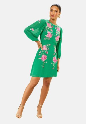 Nő zöld, térdig érő ruhában, rózsaszín virágos hímzéssel és áttetsző hosszú ujjakkal, egyik kezét a csípőjén tartva, arany magassarkú cipőben pózol.