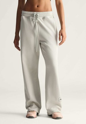 Kobieta w luźnych, jasnoszarych spodniach dresowych ze sznurkiem w pasie i bladoróżowych trampkach, stojąca na tle jednolitego jasnego tła.