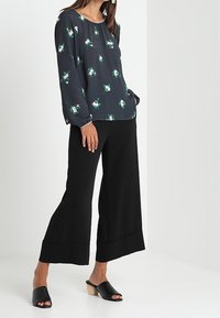 Pantaloni neri a gamba larga abbinati a una blusa floreale scura con motivi verdi e bianchi. Il modello indossa sandali neri con tacco in legno.