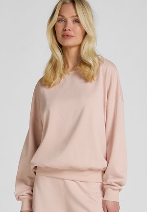 Blonde Frau trägt ein hellrosa übergroßes Sweatshirt und passende Hose vor einem einfarbigen Hintergrund.