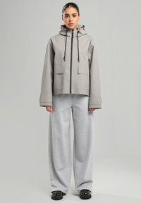 Veste imperméable gris clair avec capuche, dotée d'une fermeture éclair sur le devant, d'accents noirs et de deux poches avant. Associée à un pantalon de survêtement gris ample et à des chaussures noires.