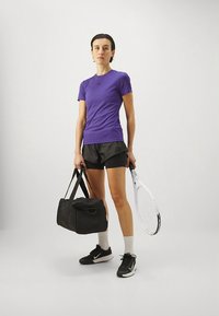 Lila sportos póló, fekete rövidnadrág, fekete sporttáska, fehér teniszütő és fekete Nike logóval ellátott sportcipők semleges háttér előtt.