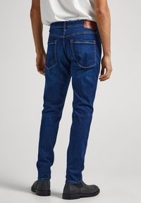 Dark blue denim jeans with a slim fit, featuring two back pockets, a brown leather tag at the waistband, and subtle stitching details.