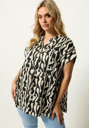 Lyshåret kvinde iført løs, kortærmet bluse med sort-hvidt abstrakt dyreprint og lyseblå jeans, stående mod en ensfarvet baggrund.