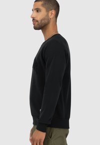 Felpa nera con maniche lunghe e collo rotondo, caratterizzata da dettagli in frange sul davanti e tessuto testurizzato. Il modello indossa pantaloni verde oliva.
