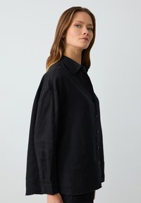 Schwarzes Leinenhemd mit kragenförmigem Ausschnitt, Knopfleiste und lockerem Schnitt. Verfügt über lange Ärmel und eine leicht strukturierte Oberfläche.