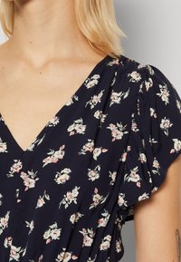 Navyblaues Blumenkleid mit V-Ausschnitt, kurzen Puffärmeln und kleinen rosa-weißen Blumen auf einem weichen, strukturierten Stoff. Details beinhalten zarte Falten.