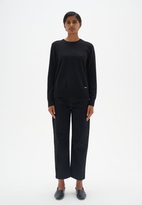 Schwarzer Pullover aus weichem Material, kombiniert mit schwarzen hoch-taillierten Jeans. Einfaches Design mit einem kleinen Markenschild auf dem Pullover.