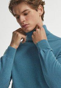 Blauer Rollkragenpullover aus weichem Stoff, mit einem enganliegenden Kragen und langen Ärmeln, samt glattem Material und minimalistischem Design.
