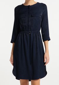 Vestido azul marino con escote redondo, cierre de botones, dos bolsillos en el pecho, cintura ajustada con un cinturón y mangas de tres cuartos. Textura suave.