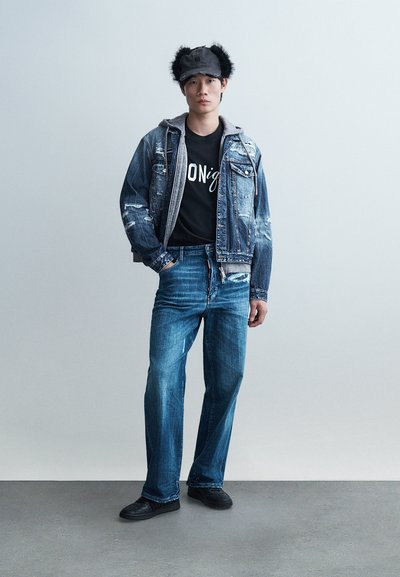 Νέος άνδρας που στέκεται με το χέρι στην τσέπη, φορώντας τζιν μπουφάν πάνω από γκρι φούτερ με κουκούλα, μαύρο γραφικό μπλουζάκι, μπλε τζιν, μαύρα παπούτσια και μαύρο καπέλο με γούνινες επενδύσεις στα αυτιά.