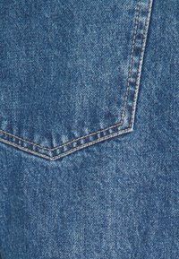 Denimstof met een blauwe tint en verschillende texturen. Zichtbare stiksels in een contrasterende kleur en een duidelijk gevormde zak.