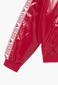 Maniche di giacca bomber rossa lucida con polsini elasticizzati, caratterizzate da una striscia con il logo "GUESS" bianco lungo il braccio. Superficie liscia, finitura lucida.