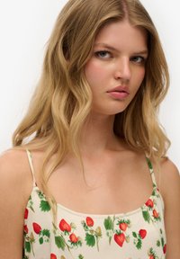 Kremfarvet top med spaghetti-stropper, der har et jordbær- og blomsterprint, glat tekstur og firkantet halsudskæring.