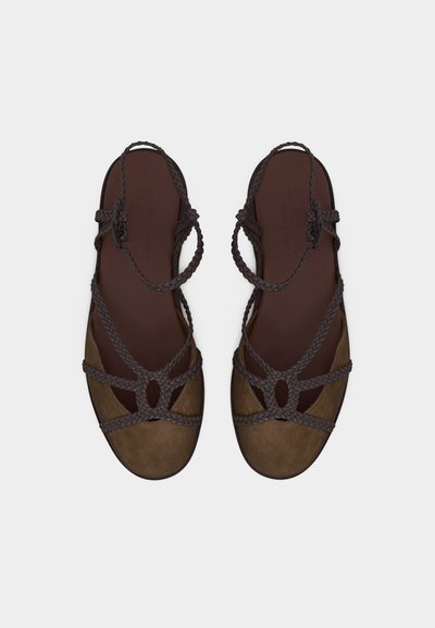Καφέ επίπεδα σανδάλια με πλεγμένα λουράκια και κλειστές στρογγυλεμένες μύτες, εμφανιζόμενα από πάνω σε λευκό φόντο.