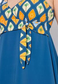 Sininen mekko, jossa on palmupuiden ja auringon kuvioita esiintyvä kuvioitu yläosa, sidottuna vyötärölle vastaavalla solmulla. Tasainen kankaan rakenne.