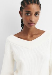 Pulover alb din tricot cu rivete, cu un decolteu lat în formă de V, având accente texturate și o croială lejeră. Materialul pare moale și ușor.