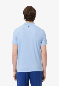 Polo shirt à manches courtes bleu clair avec un col, une texture lisse et un petit logo noir dans le dos près du col.