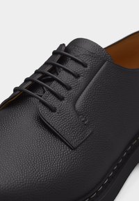 Chaussure habillée en cuir noir texturé avec laçage fermé, bout rond et couture visible sur le bord de la semelle.