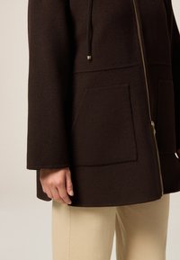 Bruine wolblendjas met een ritssluiting, brede mouwen, twee voorzakken en een gladde textuur. Gecombineerd met beige broeken.