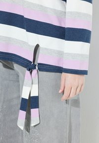 Gestreiftes Oberteil in Marineblau, Lila, Grau und Weiß aus Baumwolle, mit einem geknoteten Detail an der Seite. Kombiniert mit hellgrauen Hosen mit floralen Stickereien.