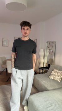 Melna īsām piedurknēm t-krekls un gaiši pelēkas sporta bikses. Fonā ir neitrālas krāsas telpa ar mūsdienīgu mēbelējumu un dekoru.