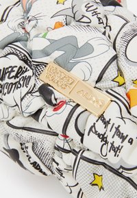 Tissu matelassé blanc avec des imprimés de bandes dessinées Looney Tunes en noir et gris, des étoiles jaunes et une étiquette métallique dorée portant la mention "Looney Tunes | ALDO."