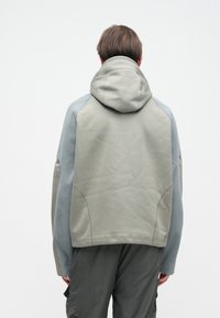 Φόρμα με κουκούλα, φτιαγμένη από ένα μείγμα γκρίζο-πράσινου υφάσματος και ανοιχτού μπλε φλίς, με χαλαρή εφαρμογή και εμφανή ραφή στους μανίκια και το σώμα.