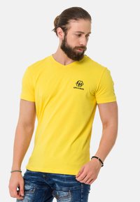 Gelbes Baumwoll-T-Shirt mit V-Ausschnitt, kurzen Ärmeln und einem schwarzen Logo auf der linken Brust. Der Stoff hat eine glatte Textur.