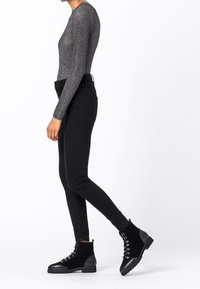 Kobieta w dopasowanej, szarej, prążkowanej bluzce z długim rękawem, czarnych, wysokich rurkach i czarnych, sznurowanych botkach sięgających do kostek, stojąca bokiem.