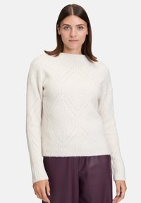Cremefarbener Rippstrickpullover mit einem strukturierten Rautenmuster, Rundhalsausschnitt und langenÄrmeln, kombiniert mit dunkelvioletten Hosen.