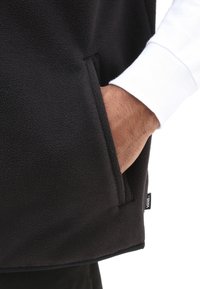 Schwarze Fleece-Weste mit Seitentasche, strukturiertem Stoff und einem kontrastierenden weißen Langarmshirt darunter. Kleines Etikett am Saum sichtbar.