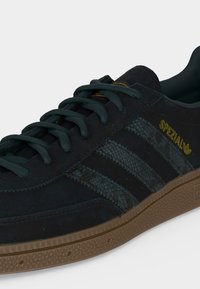 Μαύρο αθλητικό παπούτσι Adidas Spezial με υφασμένες τρεις ρίγες, καφέ σόλα και χρυσό λογότυπο στο πλάι, εμφανιζόμενο σε κοντινή λήψη.