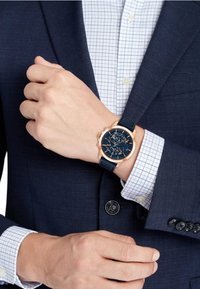 Montre marine avec un boîtier en or rose, fonctionnalités de chronographe, et un bracelet en caoutchouc bleu, portée sur une main dans un costume marine ajusté et une chemise à carreaux.