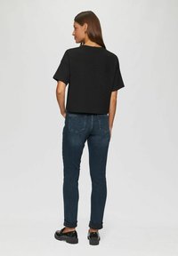 Femme de dos portant un t-shirt noir court, un jean bleu foncé retroussé et des mocassins noirs sur un fond clair.