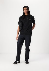 Svart kortermet t-skjorte med hvit tekst, kombinert med mørkeblå denimjeans og svarte joggesko med hvite striper. Liten svart crossbody-veske.
