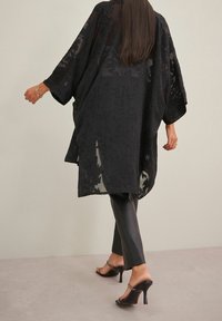 Czarny płaszcz w stylu kimono z kwiecistą koronką, szerokimi rękawami i luźnym krojem, zestawiony z dopasowanymi czarnymi spodniami i sandałami na obcasie.