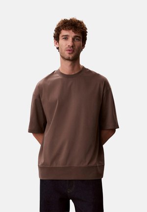 Uomo con capelli ricci che indossa una camicia marrone larga a maniche corte e pantaloni scuri su uno sfondo bianco semplice.