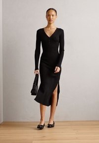 Abito lavorato a maglia nero con maniche lunghe, scollatura a V, bottoni frontali e spacco laterale. Abbinato a ballerine nere e una piccola borsa a tracolla. Texture liscia.