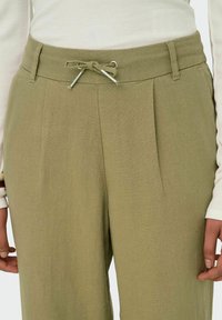 Gros plan sur un pantalon vert olive avec taille élastique, plis, et un haut à manches longues blanches partiellement visible.
