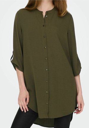 Vrouw draagt een olijfgroene blouse met lange mouwen en knopen, met opgerolde mouwen, gecombineerd met zwarte broek tegen een effen achtergrond.